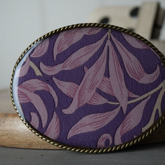 Oval-shaped purple fabric with floral pattern in a gold frame on a wooden surface.